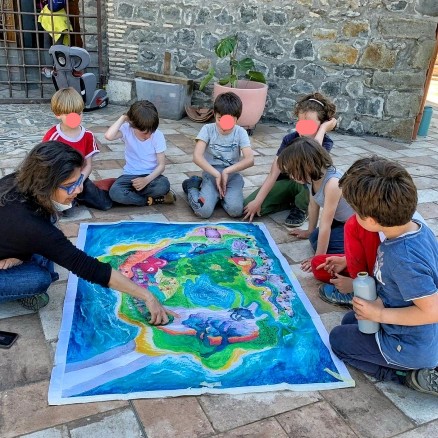 Bambini seduti a terra intorno a un dipinto insieme a una maestra; in seguito musicheranno questa immagine