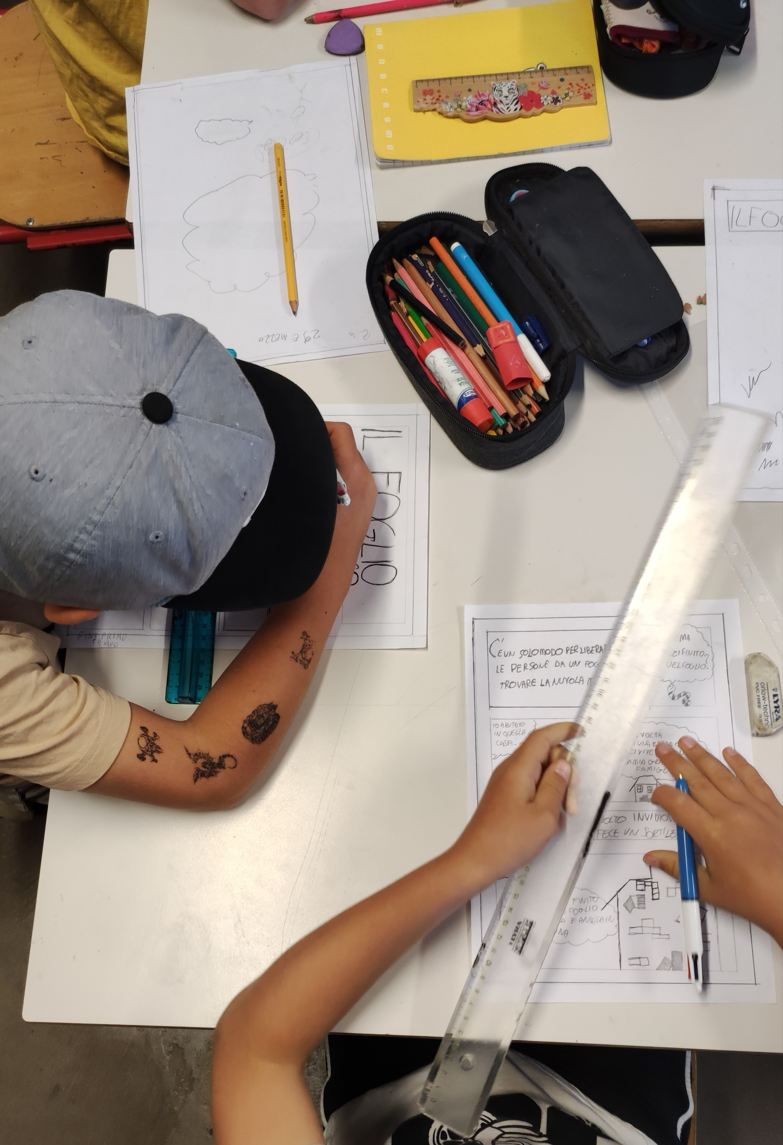 Due bambini ripresi dall'alto mentre creano il fumetto per il giornalino scolastico, con matite, penne e righelli